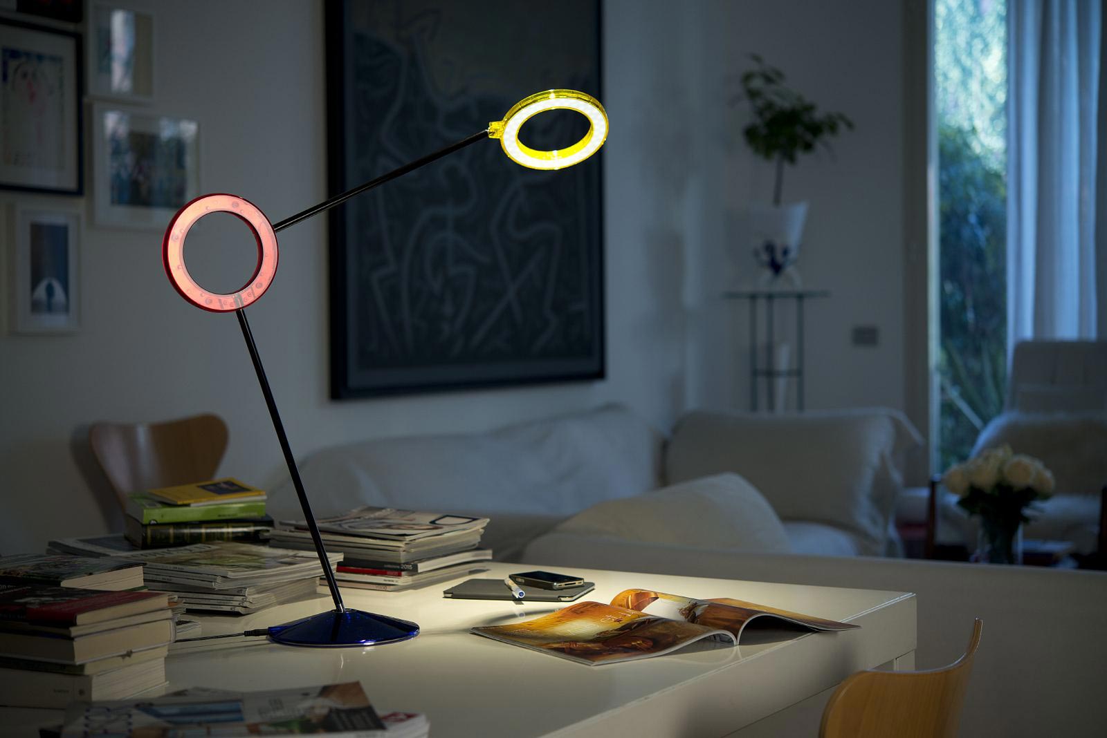 Amuleto lamp by RAMUN lit in red and yellow on a white desk in a living room